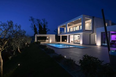 Alacakaranlıkta yüzme havuzu olan modern lüks bir evin dış görünüşü. İki katlı beyaz bir villa. Büyük camlı pencereleri, terası ve parlayan mavi suları var..