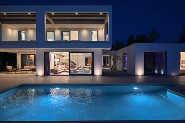 Alacakaranlıkta yüzme havuzu olan modern lüks bir evin dış görünüşü. İki katlı beyaz bir villa. Büyük camlı pencereleri, terası ve parlayan mavi suları var..