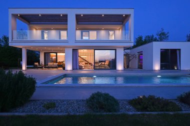 Alacakaranlıkta yüzme havuzu olan modern lüks bir evin dış görünüşü. İki katlı beyaz bir villa. Büyük camlı pencereleri, terası ve parlayan mavi suları var..