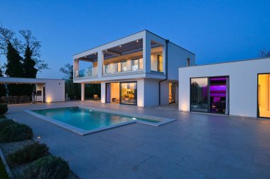 Alacakaranlıkta yüzme havuzu olan modern lüks bir evin dış görünüşü. İki katlı beyaz bir villa. Büyük camlı pencereleri, terası ve parlayan mavi suları var..