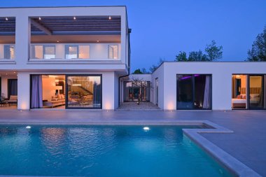 Alacakaranlıkta yüzme havuzu olan modern lüks bir evin dış görünüşü. İki katlı beyaz bir villa. Büyük camlı pencereleri, terası ve parlayan mavi suları var..