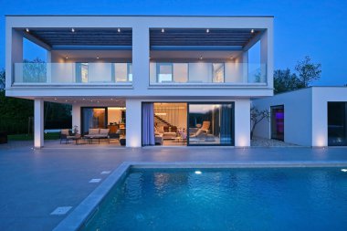 Alacakaranlıkta yüzme havuzu olan modern lüks bir evin dış görünüşü. İki katlı beyaz bir villa. Büyük camlı pencereleri, terası ve parlayan mavi suları var..
