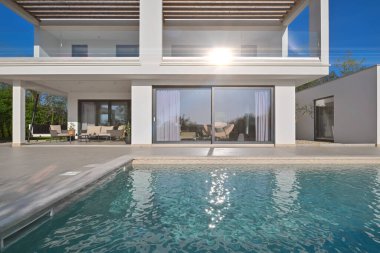 Güneşli bir günde havuzu, terası ve minimalist mimarisi olan modern lüks villa. Temiz hatlar, geniş pencereler ve açık yaşam alanları modern ev ortamları oluşturur..