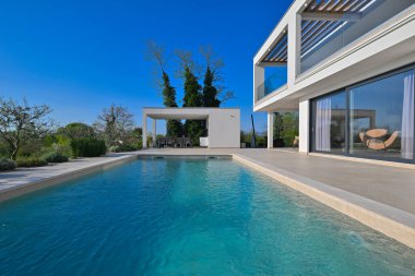 Güneşli bir günde havuzu, terası ve minimalist mimarisi olan modern lüks villa. Temiz hatlar, geniş pencereler ve açık yaşam alanları modern ev ortamları oluşturur..