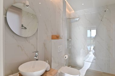 Modern, zarif banyo, beyaz mermer görünümlü fayanslar, bir damar lavabosu olan ahşap kiremit, duvara monte edilmiş tuvalet, ve parlak ortam aydınlatmasıyla aydınlatılmış cam bir duş..