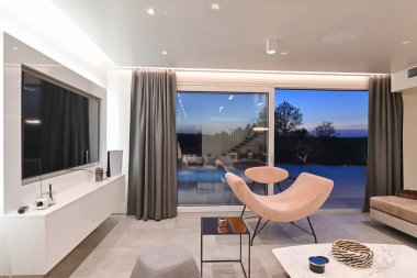 Şık modern oturma odası. Tasarımcı koltuğu, panoramik cam kapılar, günbatımı manzarası ve lüks bir ev içinde havuz başı terası..