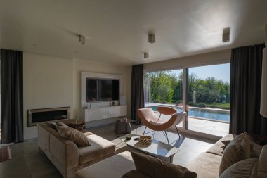 Günbatımında modern lüks oturma odası. Bej koltuklar, mermer yemek masası, parlak camlı büyük pencereler ve veranda manzaralı açık bir plan alanı. Çağdaş ev tasarımı.