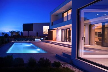 Alacakaranlıkta parlak bir yüzme havuzu olan modern lüks bir villa. Çağdaş mimari, şık tasarım ve davetkar iç ışıklar lüks bir ortam için ideal, sakin ve zarif bir akşam ortamı yaratır.