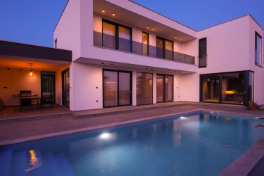 Alacakaranlıkta parlak bir yüzme havuzu olan modern lüks bir villa. Çağdaş mimari, şık tasarım ve davetkar iç ışıklar lüks bir ortam için ideal, sakin ve zarif bir akşam ortamı yaratır.