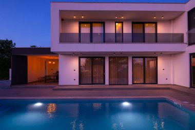 Alacakaranlıkta parlak bir yüzme havuzu olan modern lüks bir villa. Çağdaş mimari, şık tasarım ve davetkar iç ışıklar lüks bir ortam için ideal, sakin ve zarif bir akşam ortamı yaratır.