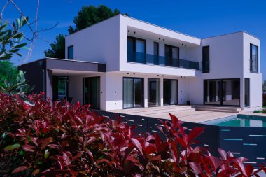 Yüzme havuzlu modern lüks villa ve mavi gökyüzünün altında temiz minimalist mimari. Geniş pencereler, teraslar ve zarif açık hava yaşam alanlarının bulunduğu çağdaş yerleşim alanı..