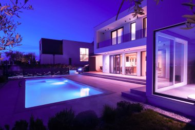 Alacakaranlıkta parlak bir yüzme havuzu olan modern lüks bir villa. Çağdaş mimari, şık tasarım ve davetkar iç ışıklar lüks bir ortam için ideal, sakin ve zarif bir akşam ortamı yaratır.