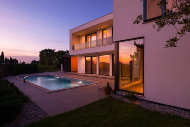 Alacakaranlıkta parlak bir yüzme havuzu olan modern lüks bir villa. Çağdaş mimari, şık tasarım ve davetkar iç ışıklar lüks bir ortam için ideal, sakin ve zarif bir akşam ortamı yaratır.