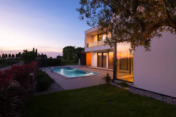 Alacakaranlıkta parlak bir yüzme havuzu olan modern lüks bir villa. Çağdaş mimari, şık tasarım ve davetkar iç ışıklar lüks bir ortam için ideal, sakin ve zarif bir akşam ortamı yaratır.