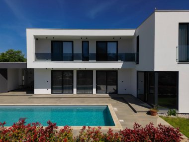 Yüzme havuzlu modern lüks villa ve mavi gökyüzünün altında temiz minimalist mimari. Geniş pencereler, teraslar ve zarif açık hava yaşam alanlarının bulunduğu çağdaş yerleşim alanı..