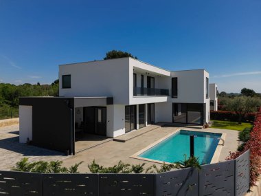 Yüzme havuzlu modern lüks villa ve mavi gökyüzünün altında temiz minimalist mimari. Geniş pencereler, teraslar ve zarif açık hava yaşam alanlarının bulunduğu çağdaş yerleşim alanı..