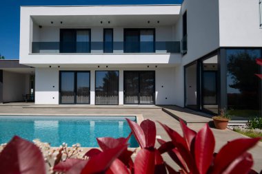 Yüzme havuzlu modern lüks villa ve mavi gökyüzünün altında temiz minimalist mimari. Geniş pencereler, teraslar ve zarif açık hava yaşam alanlarının bulunduğu çağdaş yerleşim alanı..