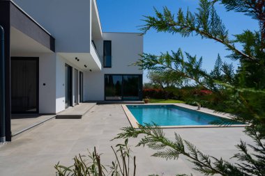 Yüzme havuzlu modern lüks villa ve mavi gökyüzünün altında temiz minimalist mimari. Geniş pencereler, teraslar ve zarif açık hava yaşam alanlarının bulunduğu çağdaş yerleşim alanı..