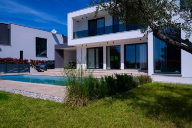 Yüzme havuzlu modern lüks villa ve mavi gökyüzünün altında temiz minimalist mimari. Geniş pencereler, teraslar ve zarif açık hava yaşam alanlarının bulunduğu çağdaş yerleşim alanı..