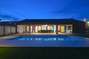 Alacakaranlıktaki lüks modern villa, sıcak iç ışıkları yansıtan sakin bir yüzme havuzu içeriyor. Davet edilen ahşap güverte, şık bir sallanan sandalyeyle tamamlanmış bir açık hava yemek alanına ev sahipliği yapıyor.