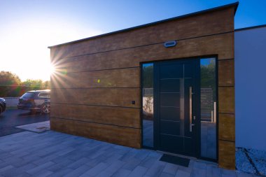 Modern bir ev girişi, şık, karanlık bir kapısı, çarpıcı ahşap bir cephesi ve kaldırımlı bir garaj yolu var. Karanlık metal geçitten geçen güneş ışığı, bu çağdaş dış evlere dinamik bir dokunuş katıyor..