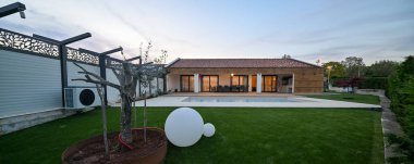 Alacakaranlıktaki lüks modern villa, sıcak iç ışıkları yansıtan sakin bir yüzme havuzu içeriyor. Davet edilen ahşap güverte, şık bir sallanan sandalyeyle tamamlanmış bir açık hava yemek alanına ev sahipliği yapıyor.