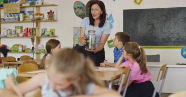 Bir sınıfta fen dersi sırasında bir grup meraklı ilkokul öğrencisine cam kavanozda teraryum bitkisini gösteren bayan öğretmen interaktif öğrenme ve keşfi teşvik ediyor. Sinema yavaşlığı