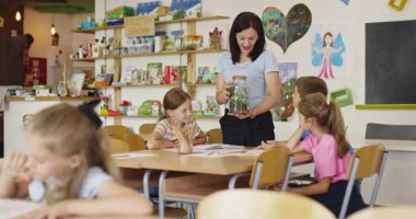 Bir sınıfta fen dersi sırasında bir grup meraklı ilkokul öğrencisine cam kavanozda teraryum bitkisini gösteren bayan öğretmen interaktif öğrenme ve keşfi teşvik ediyor. Sinema yavaşlığı