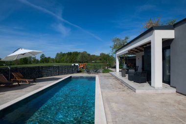 Dikdörtgen yüzme havuzu, ahşap güneş şezlongları ve mavi gökyüzüne karşı şemsiyeleri olan modern modüler evlerin arka bahçesi. Çağdaş prefabrik ev tasarımı.