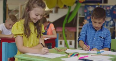 Parlak bir anaokulunda resim dersi sırasında renkli kalemlerle resim çizen yaratıcı küçük kızlara yardım eden bir bayan öğretmen..