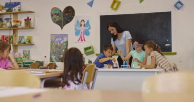 İlkokul öğrencilerine fen sınıfında deney tüpleri ve sıvılarla kimya deneyi yoluyla güvenlik gözlüğü takan bir öğrenciye rehberlik eden kadın öğretmen. Sinema yavaşlığı