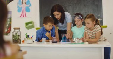 İlkokul öğrencilerine fen sınıfında deney tüpleri ve sıvılarla kimya deneyi yoluyla güvenlik gözlüğü takan bir öğrenciye rehberlik eden kadın öğretmen. Sinema yavaşlığı