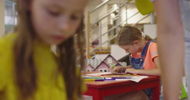 Parlak bir anaokulunda resim dersi sırasında renkli kalemlerle resim çizen yaratıcı küçük kızlara yardım eden bir bayan öğretmen..