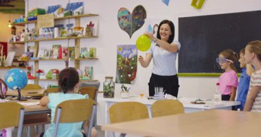 Sınıftaki heyecanlı ilkokul öğrencileri için balon ve çakmakla bilim deneyi yapan gülümseyen öğretmen, çocukların pop müzik beklentisiyle kulaklarını tıkarken,.