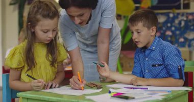 Parlak bir anaokulunda resim dersi sırasında renkli kalemlerle resim çizen yaratıcı küçük kızlara yardım eden bir bayan öğretmen..