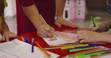 Gülümseyen bayan öğretmen iki mutlu ilkokul kızına çizimleri ve boyalarıyla yardımcı olmak için bir sınıfta yaratıcı sanat dersi sırasında masaya eğiliyor..