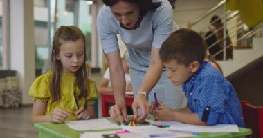 Parlak bir anaokulunda resim dersi sırasında renkli kalemlerle resim çizen yaratıcı küçük kızlara yardım eden bir bayan öğretmen..