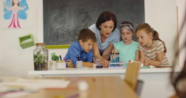 İlkokul öğrencilerine fen sınıfında deney tüpleri ve sıvılarla kimya deneyi yoluyla güvenlik gözlüğü takan bir öğrenciye rehberlik eden kadın öğretmen. Sinema yavaşlığı