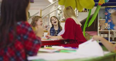 Gülümseyen bayan öğretmen iki mutlu ilkokul kızına çizimleri ve boyalarıyla yardımcı olmak için bir sınıfta yaratıcı sanat dersi sırasında masaya eğiliyor..