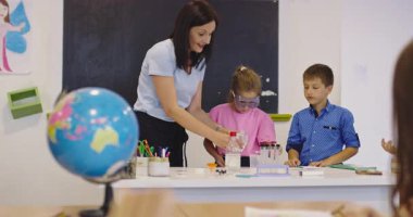 İlkokul öğrencilerine fen sınıfında deney tüpleri ve sıvılarla kimya deneyi yoluyla güvenlik gözlüğü takan bir öğrenciye rehberlik eden kadın öğretmen. Sinema yavaşlığı