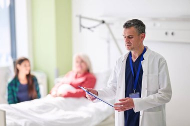 Beyaz önlüklü bir doktor, bir hastane odasında önemli bir tıbbi muayene sırasında aile üyelerinin de yardımıyla, hasta yatağında dinlenirken notlar alır..