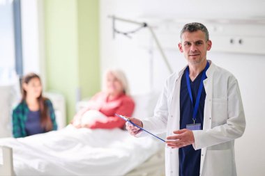 Beyaz önlüklü bir doktor elinde bir panoyla ayakta duruyor, biri hamile iki kadınla bir hastane odasında iletişim kuruyor. Atmosfer destekleyici ve profesyonel..