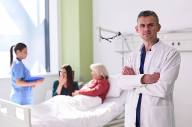 İyi aydınlatılmış bir hastane odasında, iki hemşire yatakta yatan bir hastayla ilgilenirken bir doktor kollarını kavuşturarak güvenle ayakta durur. Atmosfer profesyonel ve şefkatli hissettiriyor..