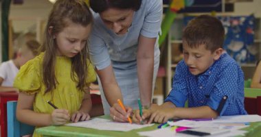 Parlak bir anaokulunda resim dersi sırasında renkli kalemlerle resim çizen yaratıcı küçük kızlara yardım eden bir bayan öğretmen..