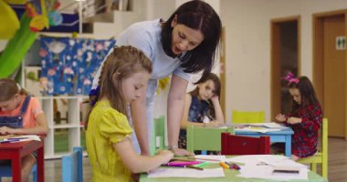 Parlak bir anaokulunda resim dersi sırasında renkli kalemlerle resim çizen yaratıcı küçük kızlara yardım eden bir bayan öğretmen..