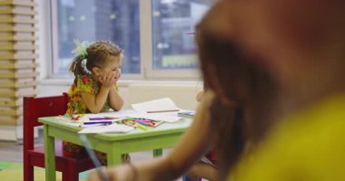 Gülümseyen bayan öğretmen iki mutlu ilkokul kızına çizimleri ve boyalarıyla yardımcı olmak için bir sınıfta yaratıcı sanat dersi sırasında masaya eğiliyor..