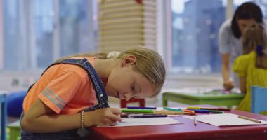 Parlak bir anaokulunda resim dersi sırasında renkli kalemlerle resim çizen yaratıcı küçük kızlara yardım eden bir bayan öğretmen..