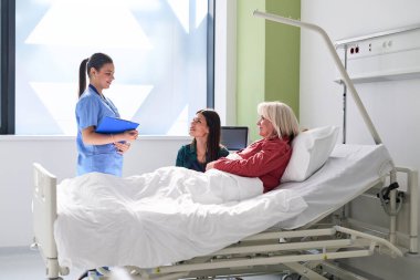 Bir hemşire hastane odasında iki kadın hastayla sohbet ediyor. Bir hasta yatakta, diğeri yanında oturuyor, ikisi de sohbete dalmış gibi görünüyor. Güneş ışığı pencereden girer..