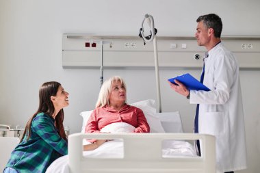 Bir doktor hastane odasında iki kadınla sağlık konularını tartışır. Bir kadın yatakta otururken diğeri dikkatle dinliyor. Ortam sakin ve şefkatli..