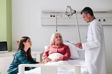 Parlak bir hastane odasında, bir doktor bir hastayla tedaviyi tartışırken, bir aile bireyi dikkatle dinler. Sahne bir anlık özen ve iletişimi yakalar..